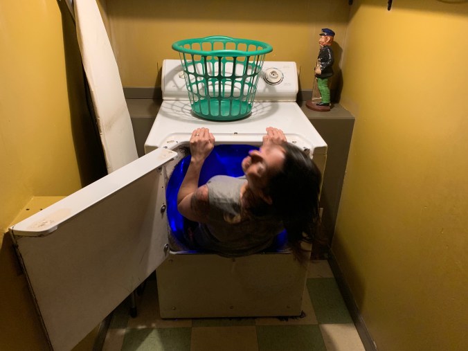 A woman crawls into a blue tunnel inside a washing machine.
