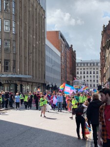 a street on a sunny day. A person in a yellow city smock leads a crowd of people waving rainbow and trans pride flags.