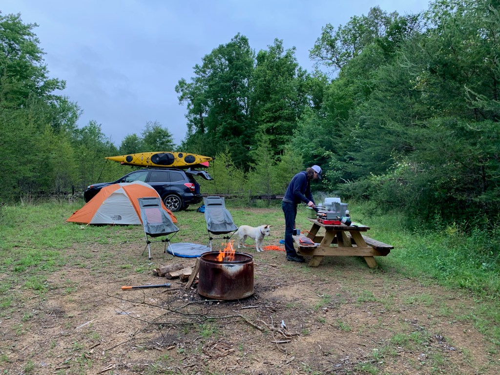 A camping scene: In the foreground, there is a campfire with two folding chairs set up nearby. In the background, a gray and orange domed tent is set up in front of a Subaru with a kayak strapped to the roof rack. On the right, a man stands over a picnic table cooking on a portable stovetop.