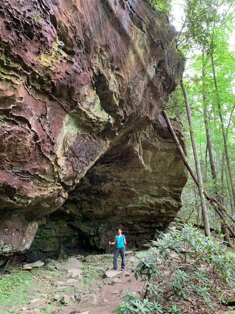 A female hiker poses in front a large rock formation multiple stories tall.